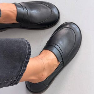 May include: A pair of black leather penny loafers featuring a rounded toe, a classic saddle strap across the vamp, and a low-profile sole. The shoes are worn with dark grey denim jeans, showcasing a smooth finish and a clean, slip-on design.
