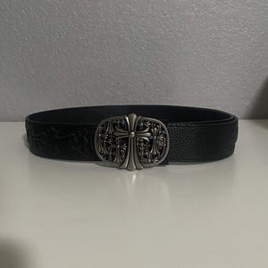 May include: Black leather belt with a silver-colored cross buckle. The belt has a textured surface and a decorative pattern. The buckle is oval-shaped with a detailed cross design. A stylish accessory.