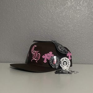 May include: A dark brown baseball cap featuring pink embroidered gothic lettering and cross symbols on the front panel. The cap includes several attached circular brand tags with black and white graphics, including text that reads Chrome Hearts.
