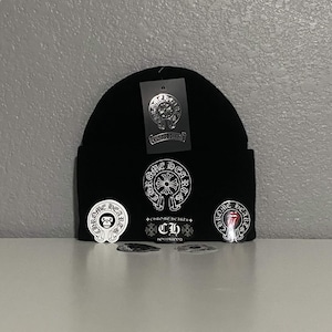 May include: A black knit beanie featuring a white embroidered Chrome Hearts logo on the front, accompanied by a matching black tag and three circular stickers with various graphic designs including a monkey and a tongue logo attached to the front.