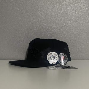 May include: A black baseball cap featuring tonal embroidered gothic lettering and cross motifs on the side. Several circular stickers with the text CHROME HEARTS and graphic designs are attached to the front panel and placed near the base.