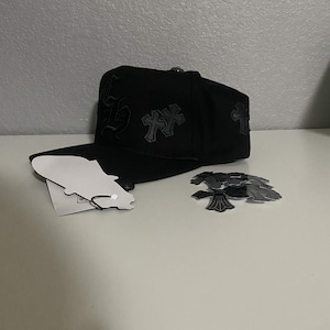 May include: A black baseball cap featuring tonal embroidered cross motifs and gothic lettering on the side panels. The cap includes a flat brim, a silver-toned button on the crown, and a white paper hang tag attached with a black cord.