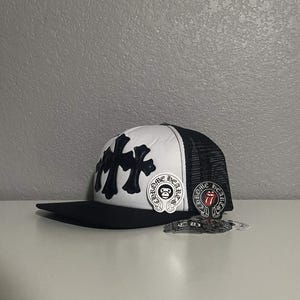 May include: A black and white trucker hat featuring a black flat brim and a white front panel adorned with black cross appliqués. The side panels are made of black mesh. Two circular stickers are attached, one with a skull and one with a red tongue logo.