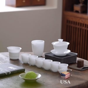 Può includere: Un set da tè in porcellana bianca composto da un gaiwan con coperchio su piattino, una brocca alta per versare e sette piccole tazze da tè. Il set è disposto su un vassoio rettangolare grigio scuro, con un piccolo oggetto verde in una ciotola.