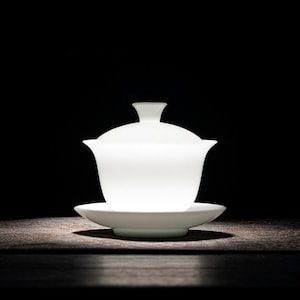 May include: A white ceramic tea set with a lidded teapot and a saucer. The set is on a dark wooden surface.