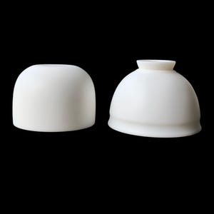 May include: Two white ceramic lamp shades against a black backdrop. One is a rounded dome, the other a rounded dome with a small, cylindrical top. The shades are smooth and opaque, suitable for a table lamp.