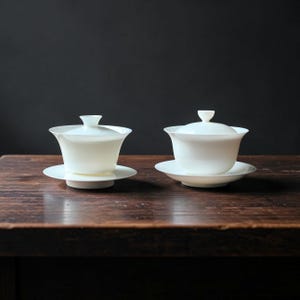 May include: Two white porcelain tea sets, each with a lidded cup and saucer, are arranged on a dark wooden surface. The tea sets have a classic design with a smooth, glossy finish, and are set against a dark background.