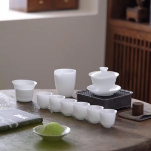 May include: A white porcelain tea set, including a lidded gaiwan, a pitcher, and several small cups, arranged on a wooden table. A small tray holds the gaiwan. A small plate with a green item is in the foreground.