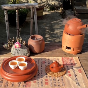 Puede incluir: Un juego de té tradicional con una tetera, tazas y un calentador de terracota. El juego incluye una bandeja redonda con tres tazas, una tetera pequeña sobre una esterilla tejida y un tarro de cerámica. El calentador de té emite vapor.