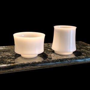 May include: Two white, opaque glass cups with a flared design sit on a dark, speckled tray. One cup is shorter and wider, while the other is taller and narrower. The cups have a simple, elegant shape, suitable for beverages.