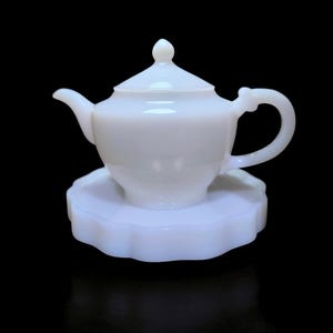 May include: A white, opaque teapot with a matching lid and a curved handle. The teapot sits on a scalloped, white saucer. The set is made of a glossy material and is set against a black background.