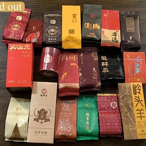 May include: An assortment of tea packages in various colors, including red, yellow, green, and brown. The packages feature Chinese characters and designs, with some labeled "Sold out". A small, red metal container is also visible.