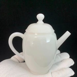 Könnte beinhalten: Eine weiße Keramik-Teekanne mit gebogenem Griff, Ausguss und Deckel mit kleinem Knauf. Die Teekanne hat eine glänzende Oberfläche. Schwarzer Hintergrund.