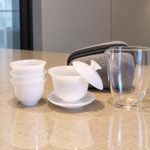 以下が含まれることがあります： 積み重ねられた3つの小さな白い磁器のティーカップ、蓋と受け皿付きの白い磁器の蓋碗、透明なガラス製のピッチャー、そしてグレーのファスナー付きキャリングケースのセットが、滑らかな表面の上に配置されています。