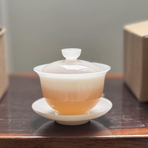 Könnte beinhalten: Eine weiß-orangefarbene Teetasse mit Deckel und Untertasse. Die Teetasse ist mit Tee gefüllt und steht auf einer dunklen Holzoberfläche. Im Hintergrund befinden sich zwei Pappkartons.