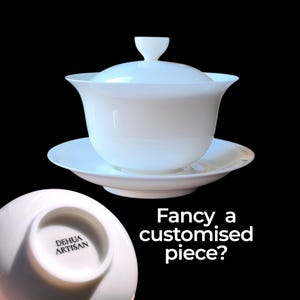May include: A white porcelain tea set, comprising a lidded cup and saucer, set against a black backdrop. The cup features a rounded form with a flared rim and a small lid handle. The saucer is circular. Text on the saucer reads "DEHUA ARTISAN".