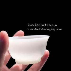 May include: A white teacup with a comfortable sipping size, as stated in the text. The teacup is held in a hand against a black background. The text on the image says "70ml (2.3 oz) Teacup, a comfortable sipping size."