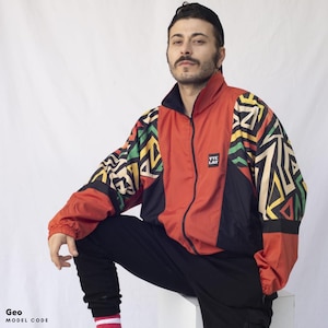 Op de afbeelding: Een rood, zwart, geel en groen gepatroneerd jack met een ritssluiting. Het jack heeft een zwart logo met de tekst "VTE LAB" op de linkerborst.