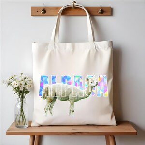 Op de afbeelding: Een witte canvas tote bag met een kleurrijk geometrisch ontwerp dat het woord "ALPACA" spelt. Een wit alpaca staat voor het woord.