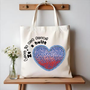 May include: White canvas tote bag with a blue heart design and the text "Love is not found, It's built".