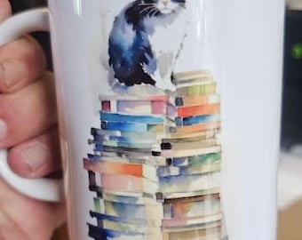 May include: White ceramic mug featuring a watercolor illustration of a black and white cat sitting atop a stack of colorful books. The mug has a white handle and a glossy finish.
