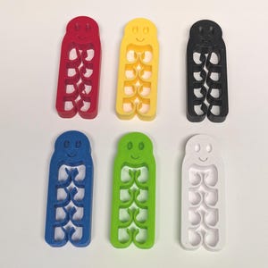Puede incluir: Seis cortadores de galletas con forma de gusano sonriente en rojo, amarillo, negro, azul, verde y blanco. Cada cortador tiene una serie de recortes curvos y una cara sonriente en la parte superior. Probablemente se utilizan para hornear o manualidades.