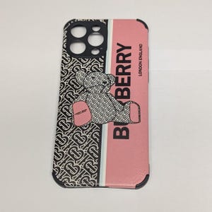 Puede incluir: Funda de teléfono rosa y negra con un diseño de oso de peluche. La funda presenta la palabra "Burberry" en letras grandes y en negrita, y las palabras "London England". El lado izquierdo de la funda tiene un patrón repetido de la letra "B".