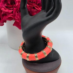 Op de afbeelding: Een scharnierende armband met afwisselende segmenten van fel koraalkleurig emaille en gepolijst goudkleurig metaal. Het ontwerp bestaat uit gladde, afgeronde koraalkleurige secties gescheiden door geribbelde goudkleurige banden voor een gesegmenteerde look.