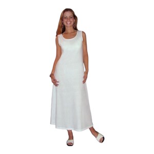 May include: A sleeveless, white maxi dress with a scoop neckline. The dress has a flowing, A-line silhouette and is paired with white sandals. The fabric appears to have a subtle texture. The model is smiling.