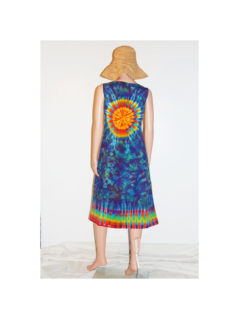 May include: A sleeveless tie-dye dress with a vibrant sunburst design on the back, transitioning to a rainbow hem. The dress is displayed on a mannequin, paired with a straw sun hat. The dress is a knee-length style.