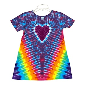 May include: A tie-dye dress with a heart-shaped design in the center. The dress features a vibrant rainbow pattern with shades of purple, blue, red, orange, yellow, and green. The short-sleeved dress is hanging on a white hanger.