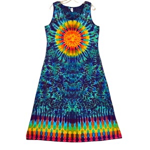 May include: Sleeveless tie-dye dress with a vibrant, colorful design. The dress features a circular pattern in the center, transitioning to shades of blue and green. The bottom hem has a rainbow stripe. The dress is hanging on a white hanger.