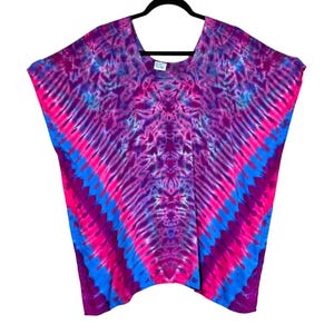 Könnte beinhalten: Ein Batik-Top in Pink-, Lila- und Blautönen, das an einem schwarzen Kleiderbügel hängt. Das Oberteil hat einen V-Ausschnitt und eine fließende, lockere Passform. Das Muster ist symmetrisch, mit Streifen, die von der Mitte ausgehen.