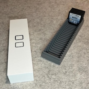 May include: A white rectangular case with two black rectangles and a gray game cartridge holder. The holder contains several black game cartridges, one labeled "Harry Potter".