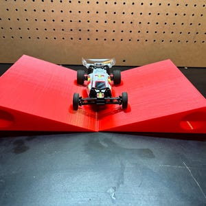 Peut inclure: Une rampe en plastique rouge avec deux trous de chaque côté, conçue pour une voiture jouet. Une voiture jouet blanche avec des roues noires est positionnée sur la rampe.