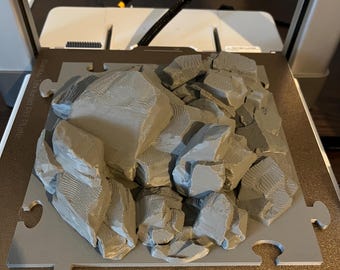 Op de afbeelding: Een 3D-geprint model van een rotsachtig landschap, in grijstinten, op een puzzelvormige basis. Het model heeft gedetailleerde texturen en rotsen van verschillende grootte, wat een miniatuurterrein suggereert. De basis heeft de tekst "PLA/ABS/PETG".