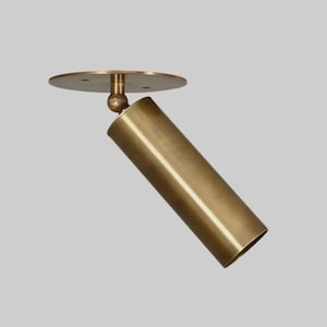 May include: A brass-colored, cylindrical spotlight fixture. The light is mounted on a circular base and connected by a ball joint, allowing for directional adjustment. The fixture is designed for ceiling mounting and has a modern aesthetic.