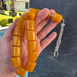 May include: A strand of amber-coloured prayer beads with rectangular beads. The beads are a warm, translucent yellow-orange hue. The strand is held in a hand, with a silver tassel and decorative end piece.