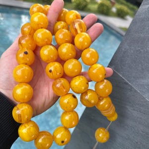 May include: A necklace or prayer beads made of large, spherical amber beads. The beads are a rich, golden-yellow colour with darker flecks. The beads are strung together, forming a continuous loop. The background is blurred, suggesting an outdoor setting.