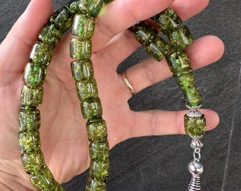 Può includere: Una collana di perline di preghiera verde e oro. Le perline rettangolari e traslucide hanno scaglie d'oro. La collana ha un nappa d'argento e un elemento terminale decorativo. Le perline sono tenute in una mano.