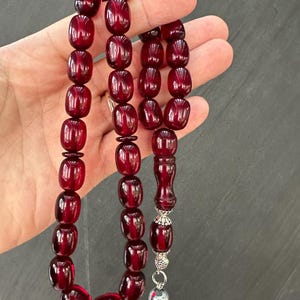 Puede incluir: Un rosario o cuentas de oración de ámbar rojo con un borla de plata y una cuenta de plata en el extremo. Las cuentas son de forma ovalada y tienen una superficie ligeramente texturizada.