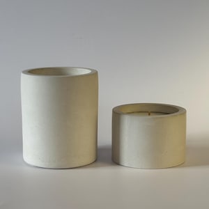 May include: Two cylindrical, off-white concrete candle holders. One is tall and empty, the other is shorter and contains a candle. The candle has a visible wick and a light yellow wax. The holders have a smooth, matte finish.