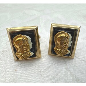 May include: A pair of rectangular cufflinks featuring a gold-toned relief of a helmeted figure in profile set against a black background. The metallic frame is gold-toned, and the design depicts a warrior with a beard wearing a classic helmet.