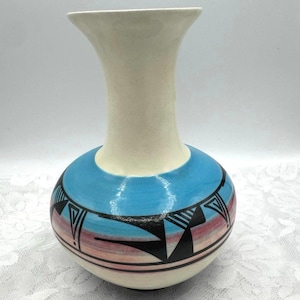 May include: A ceramic vase with a cream-colored neck and a round, blue base. The base features a geometric design in black, red, and pink. The vase has a flared opening and a smooth finish. The vase is approximately 8 inches tall.