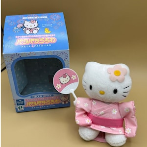 May include: A Hello Kitty plush toy wearing a pink kimono and holding a matching fan. The fan and kimono are decorated with white flowers. A blue box with Japanese text and Hello Kitty graphics is in the background.