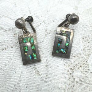 May include: A pair of rectangular silver-toned clip-on earrings featuring a central Greek key motif inlaid with iridescent green and blue abalone shell fragments. The earrings have a polished metal frame and a rounded screw-back closure mechanism.