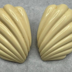 May include: Two cream-colored, shell-shaped earrings. Each earring has a ribbed design, with a glossy finish. The earrings are set against a light gray background. The earrings are a vintage style.