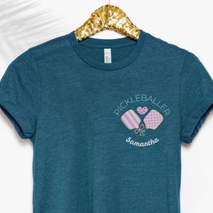 May include: Teal blue t-shirt with a white graphic of two pink and white pickleball paddles with the text "Pickleballer Samantha" in white.