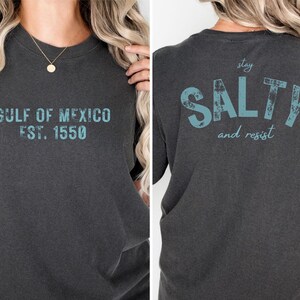 May include: A charcoal gray short-sleeve t-shirt featuring teal distressed text. The front displays 'GULF OF MEXICO EST. 1550' and the back features the text 'stay SALTY and resist' in a large, bold, weathered font style.