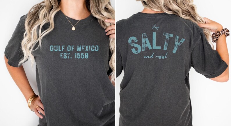 May include: A charcoal gray short-sleeve t-shirt featuring teal distressed text. The front displays 'GULF OF MEXICO EST. 1550' and the back features the text 'stay SALTY and resist' in a large, bold, weathered font style.
